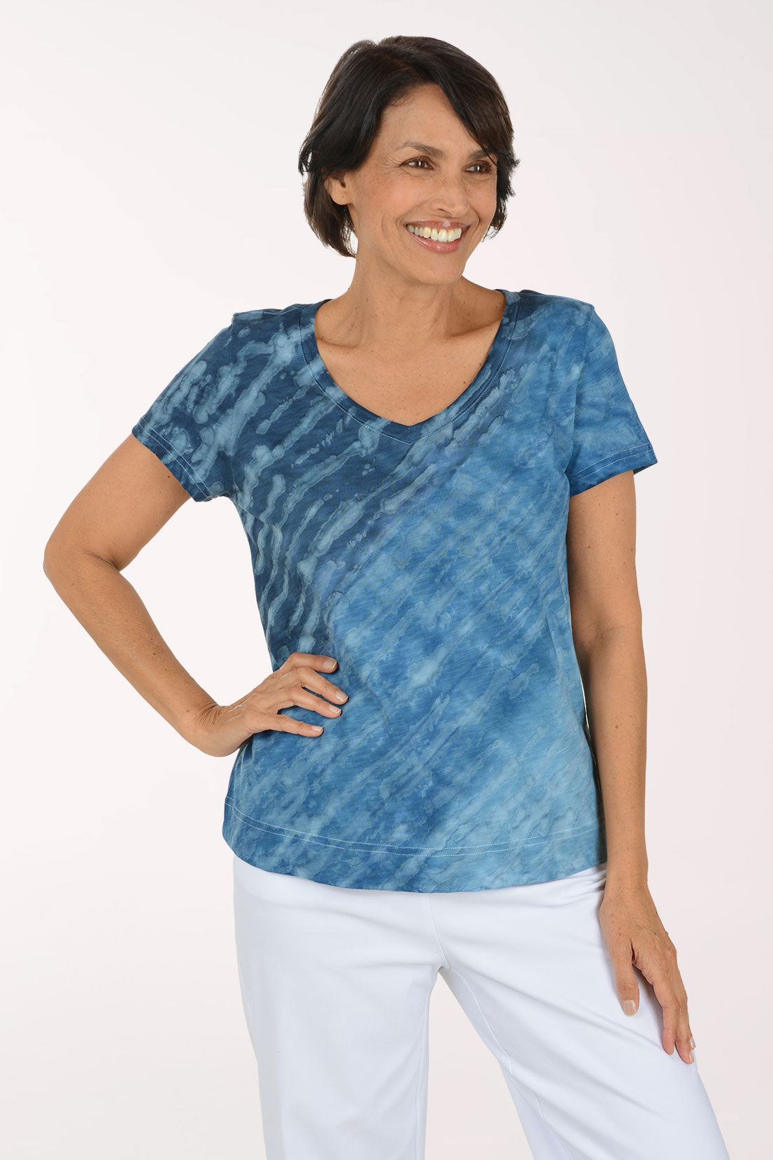 A woman modeling a blue tie-dye short-sleeve top with a V-neck, paired with white pants, against a plain white studio background.