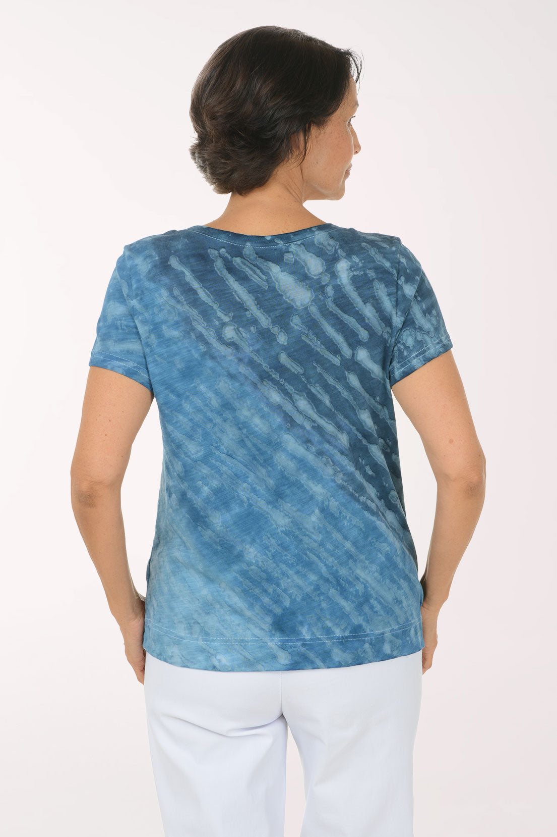 A woman modeling a blue tie-dye short-sleeve top with a V-neck, paired with white pants, against a plain white studio background.