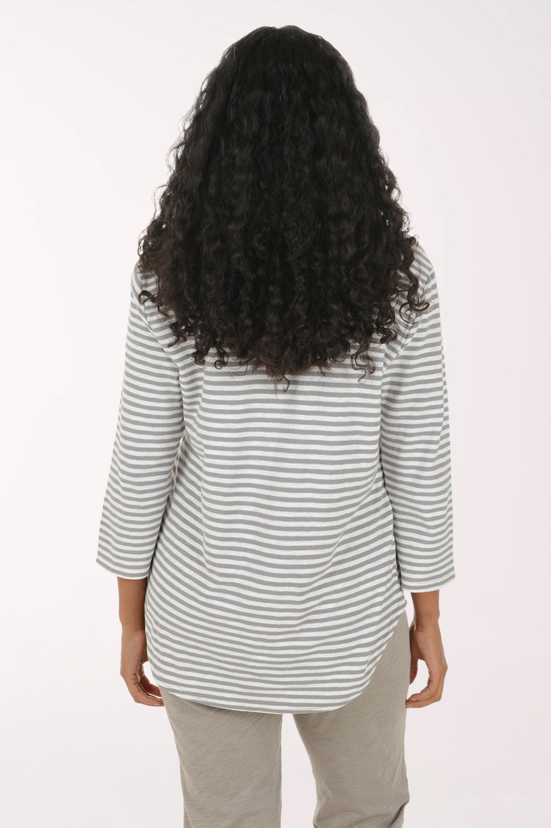 A smiling woman with long curly black hair models a gray and white striped button-up shirt with three-quarter sleeves, paired with heather gray pants, against a plain white studio background.