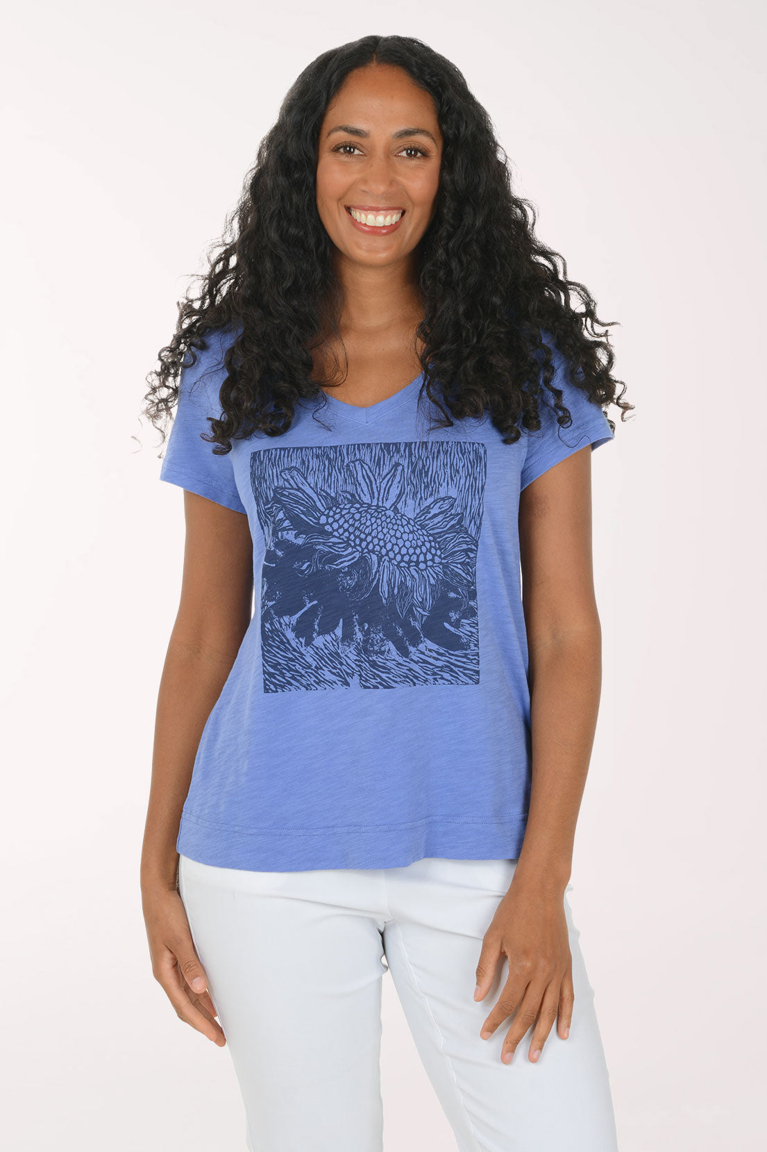 A woman with long, curly black hair wearing a heathered blue V-neck t-shirt with a dark blue sunflower graphic on the front, paired with white pants, standing against a plain white studio background.