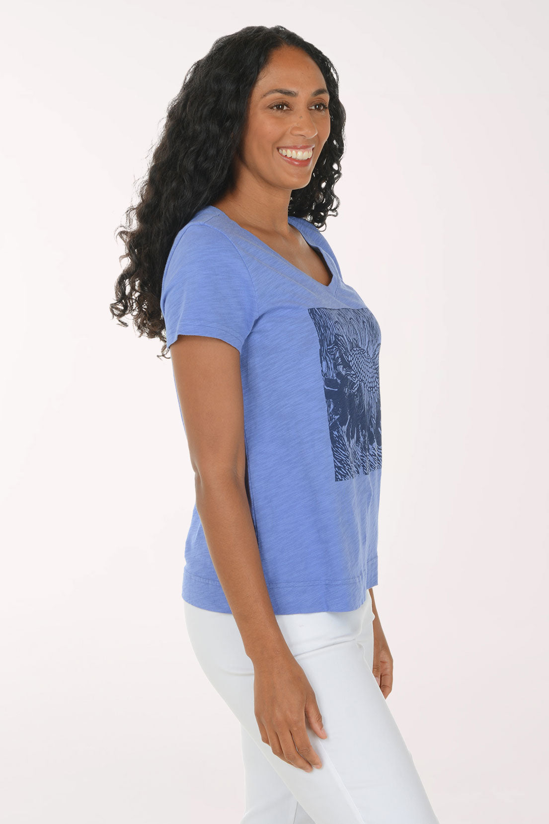 A woman with long, curly black hair wearing a heathered blue V-neck t-shirt with a dark blue sunflower graphic on the front, paired with white pants, standing against a plain white studio background.