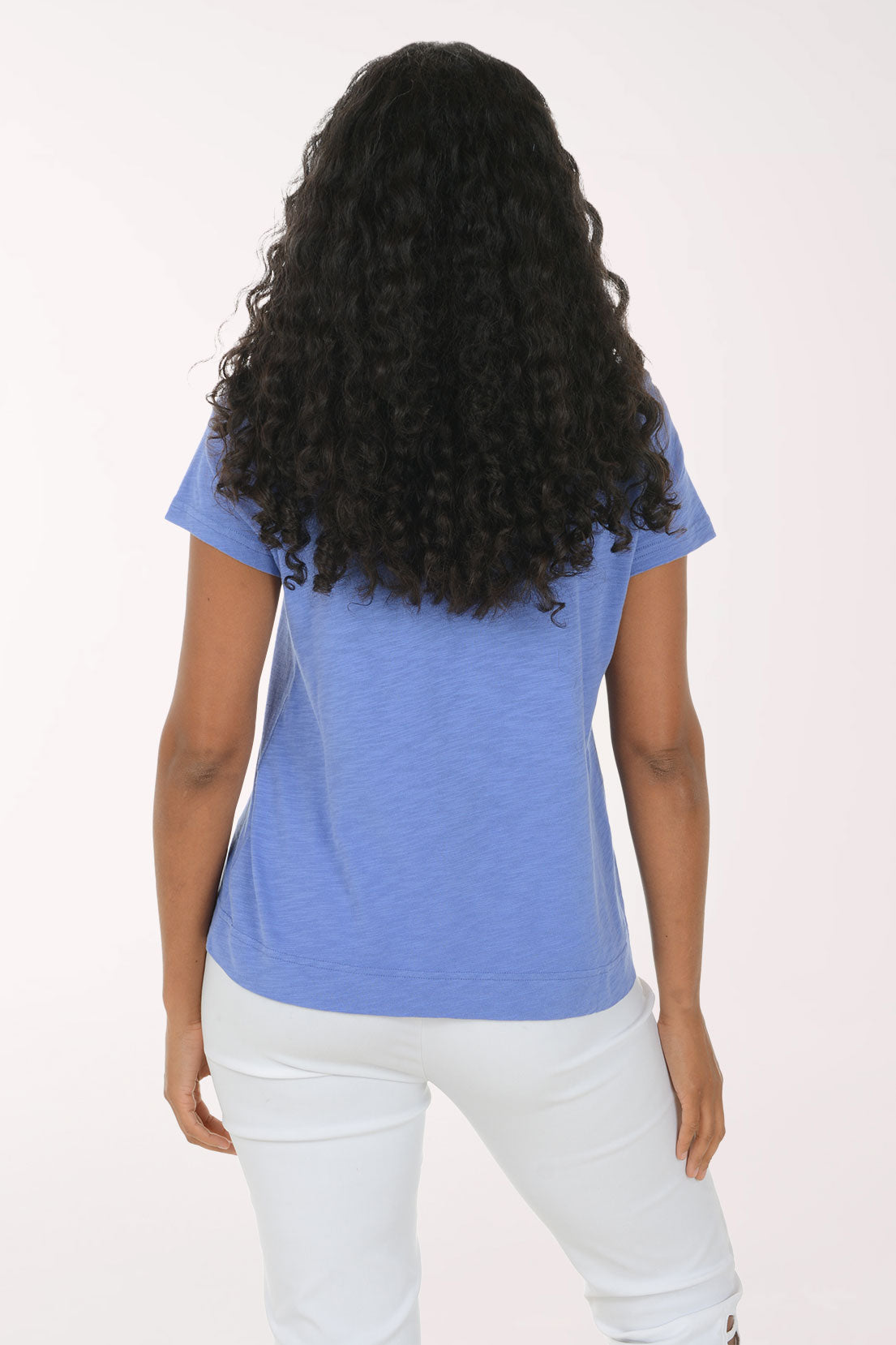 A woman with long, curly black hair wearing a heathered blue V-neck t-shirt with a dark blue sunflower graphic on the front, paired with white pants, standing against a plain white studio background.