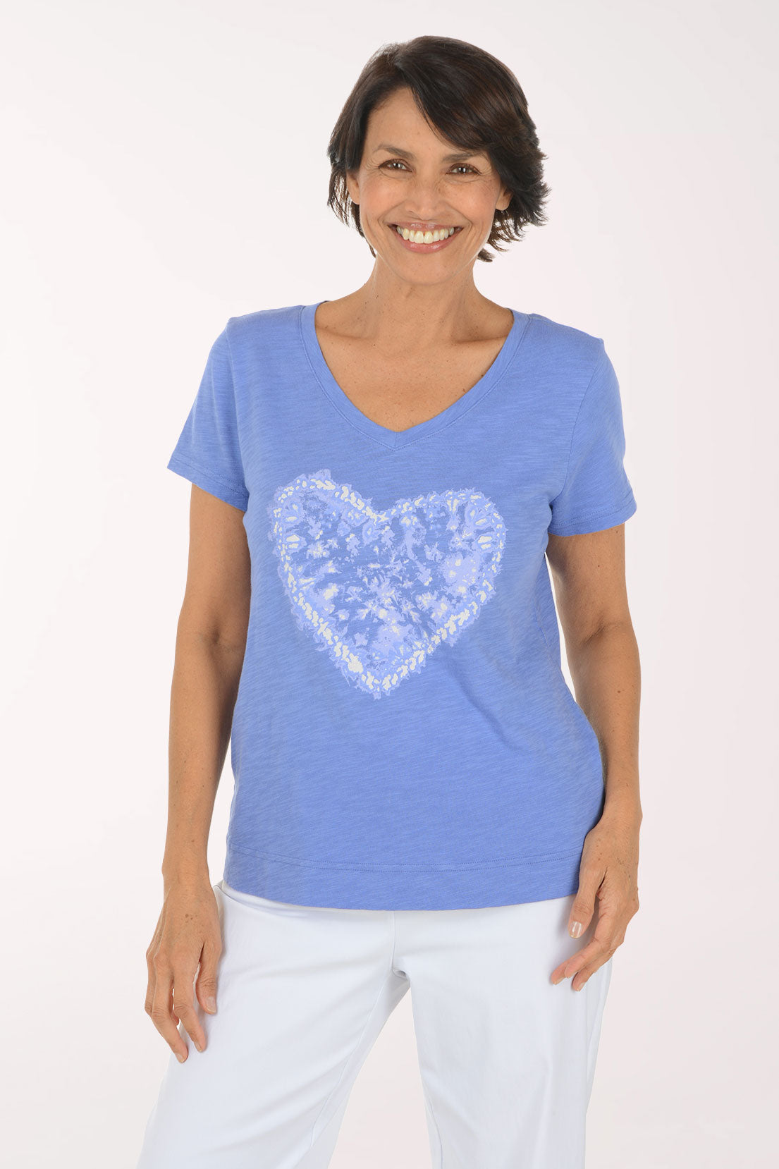 A smiling woman wearing a heathered periwinkle blue V-neck t-shirt with a white heart-shaped graphic on the front, paired with white pants, against a plain white studio background.