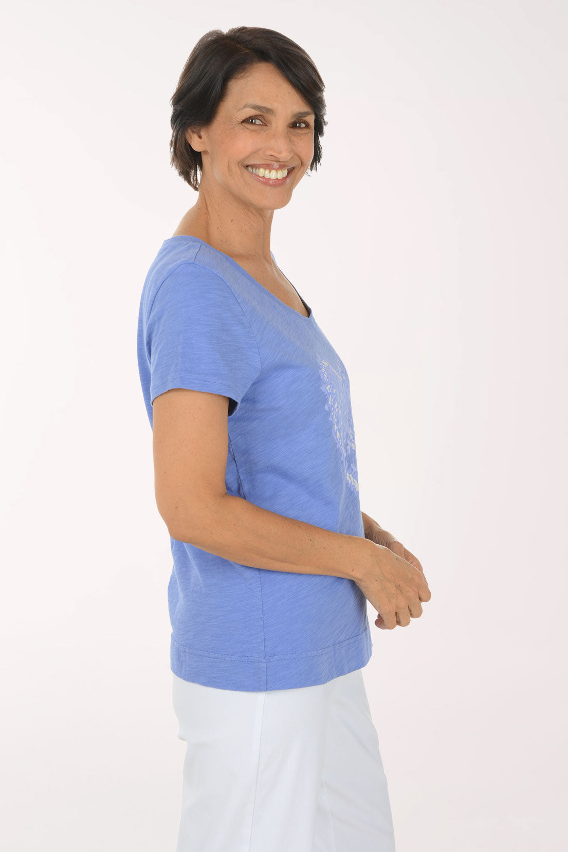 A smiling woman wearing a heathered periwinkle blue V-neck t-shirt with a white heart-shaped graphic on the front, paired with white pants, against a plain white studio background.