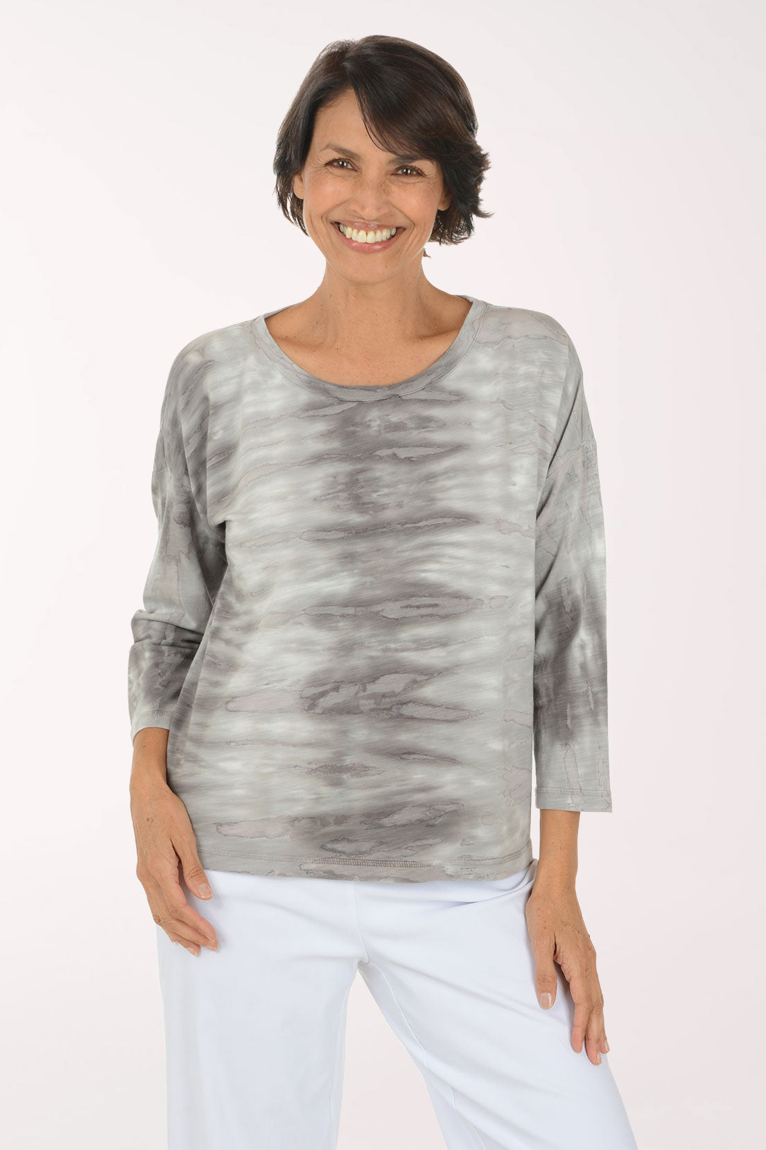 A smiling woman wearing a gray and white tie-dye long-sleeve top with white pants, standing against a plain light background.