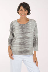 A smiling woman wearing a gray and white tie-dye long-sleeve top with white pants, standing against a plain light background.
