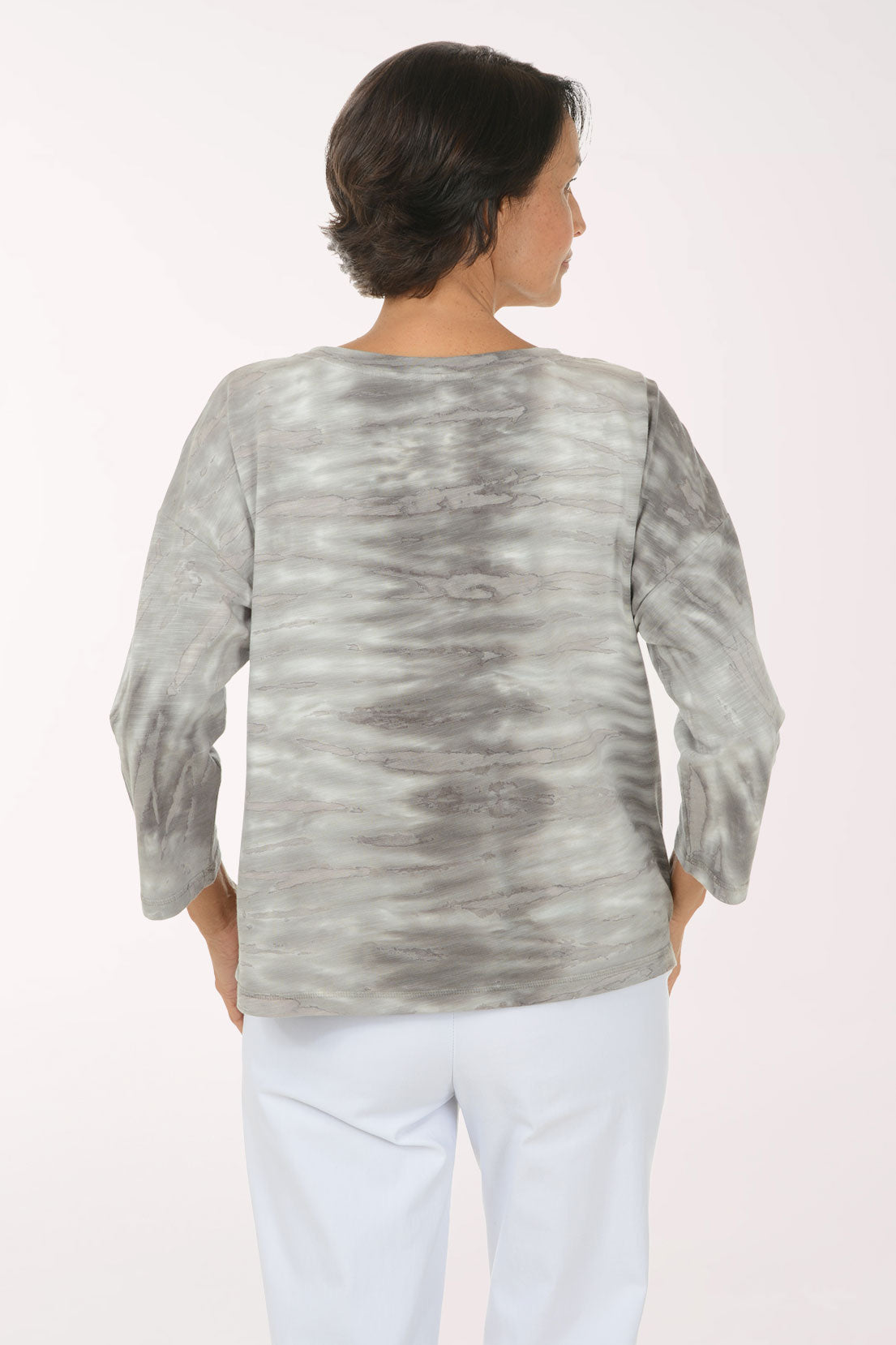 A smiling woman wearing a gray and white tie-dye long-sleeve top with white pants, standing against a plain light background.