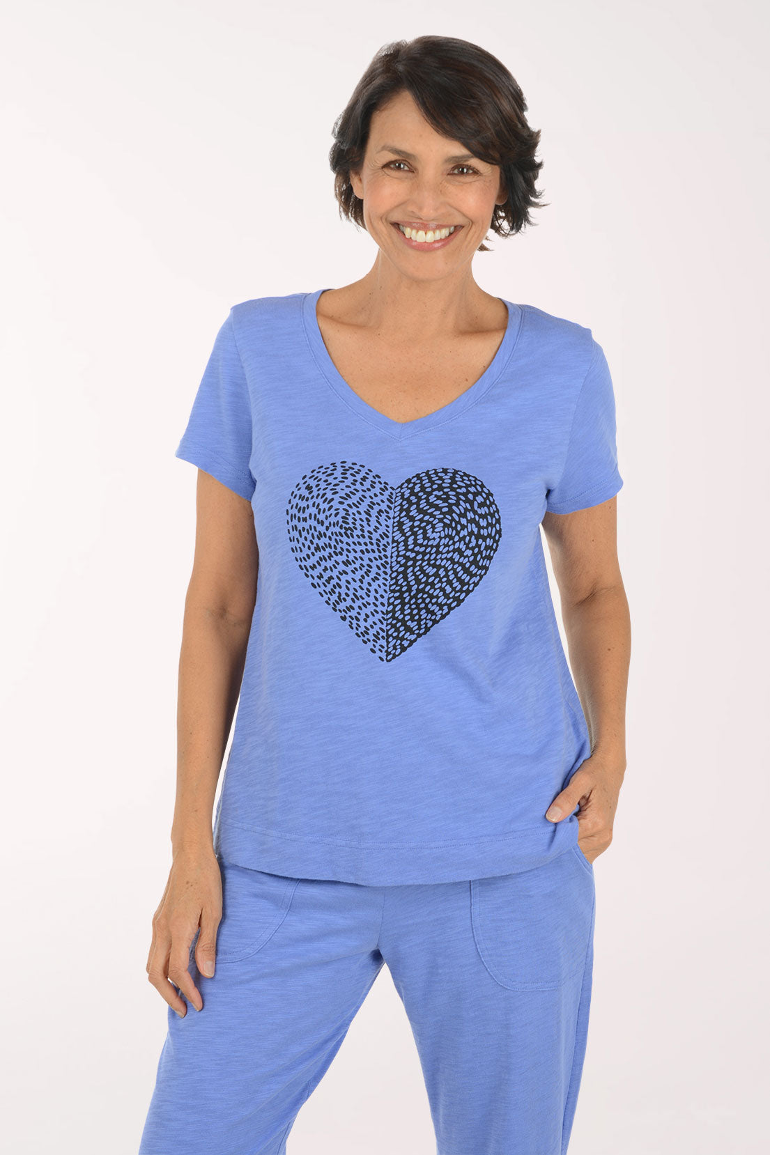 A smiling woman modeling a heathered blue V-neck short-sleeve top with a black dotted heart graphic on the chest, paired with matching blue pants, against a plain white studio background.
