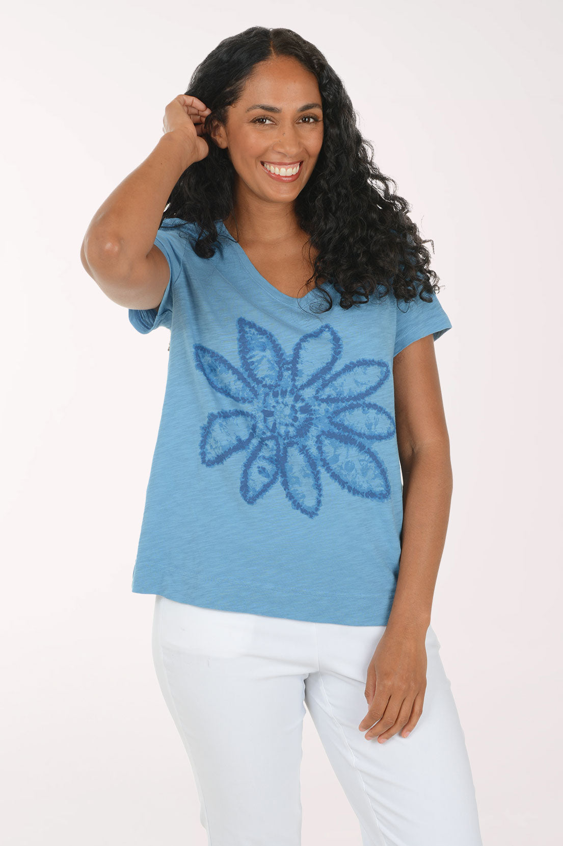 A woman wearing a heathered light blue t-shirt with a textured blue flower graphic on the front, paired with white pants, against a plain light background.