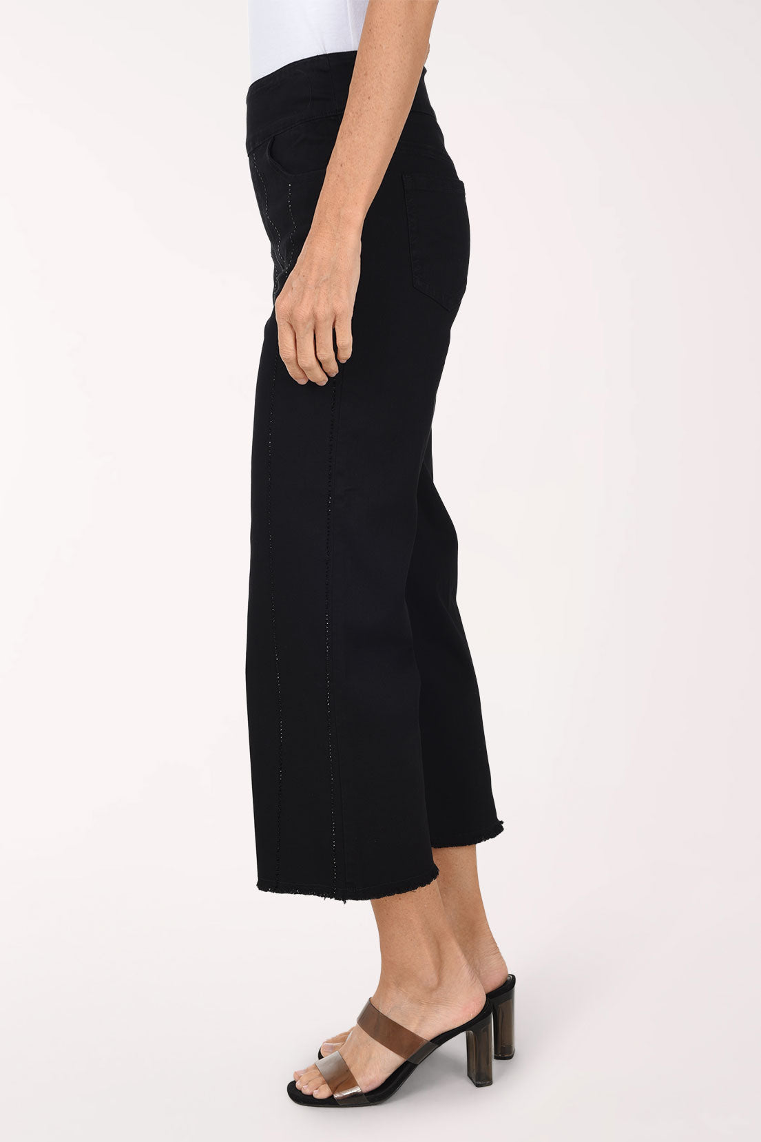 A side view of black wide-leg denim pants with vertical rhinestone stripes and a frayed hem, worn with clear-strap heeled sandals.