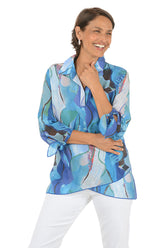 Woman wearing a colorful patterned blouse with white pants on a white background.