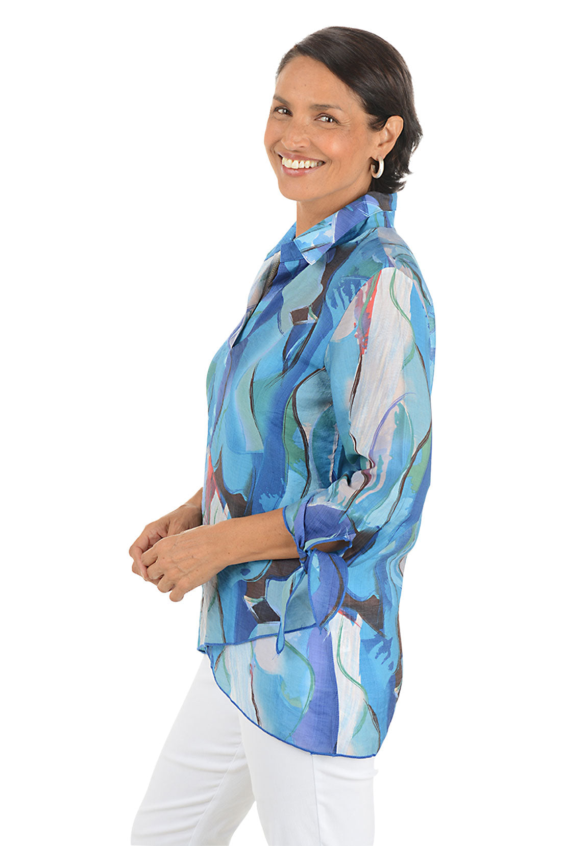 Woman wearing a colorful patterned blouse with white pants on a white background. Side shot.