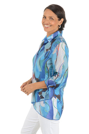 Woman wearing a colorful patterned blouse with white pants on a white background. Side shot.