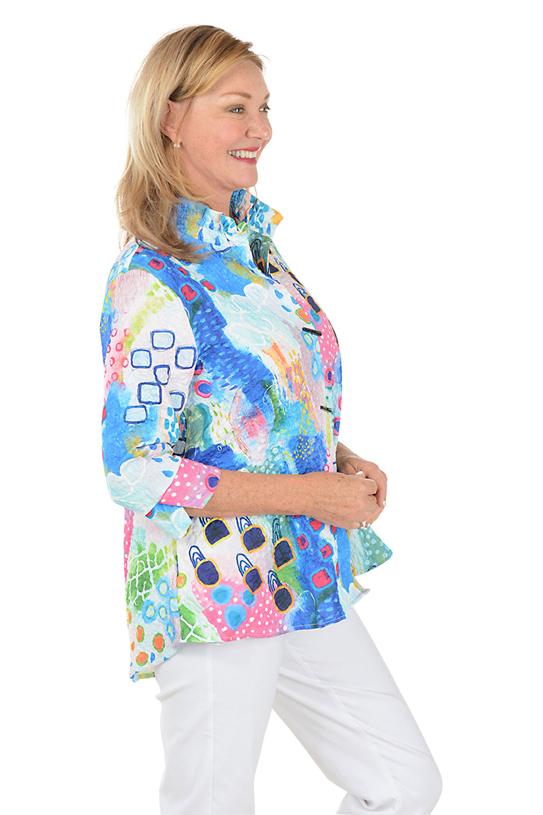 Woman wearing a colorful patterned blouse with white pants on a white background. Side shot.