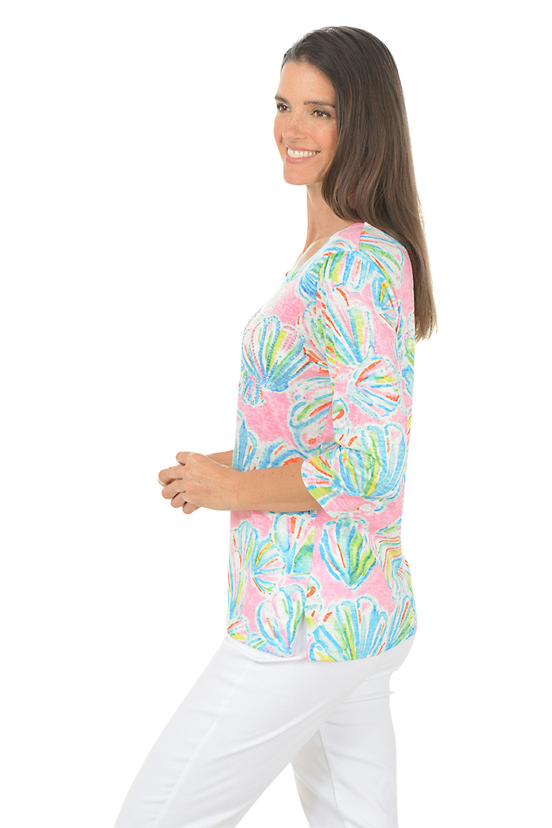 Woman wearing a colorful patterned top with white pants on a white background. Side shot.
