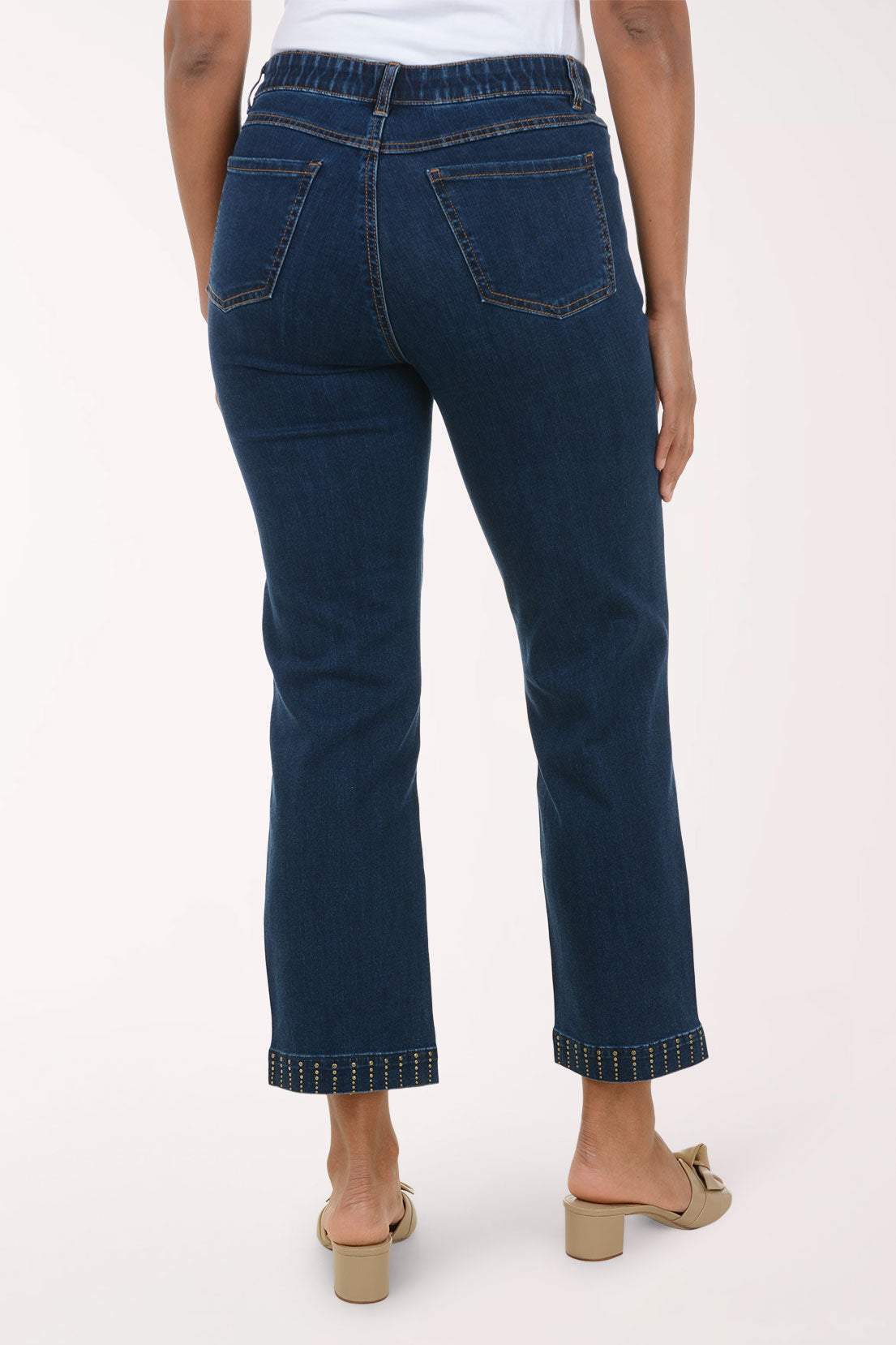 Rear view of a person wearing dark blue cropped jeans with rolled hems and gold-toned contrast stitching, paired with beige mule sandals against a plain light background.