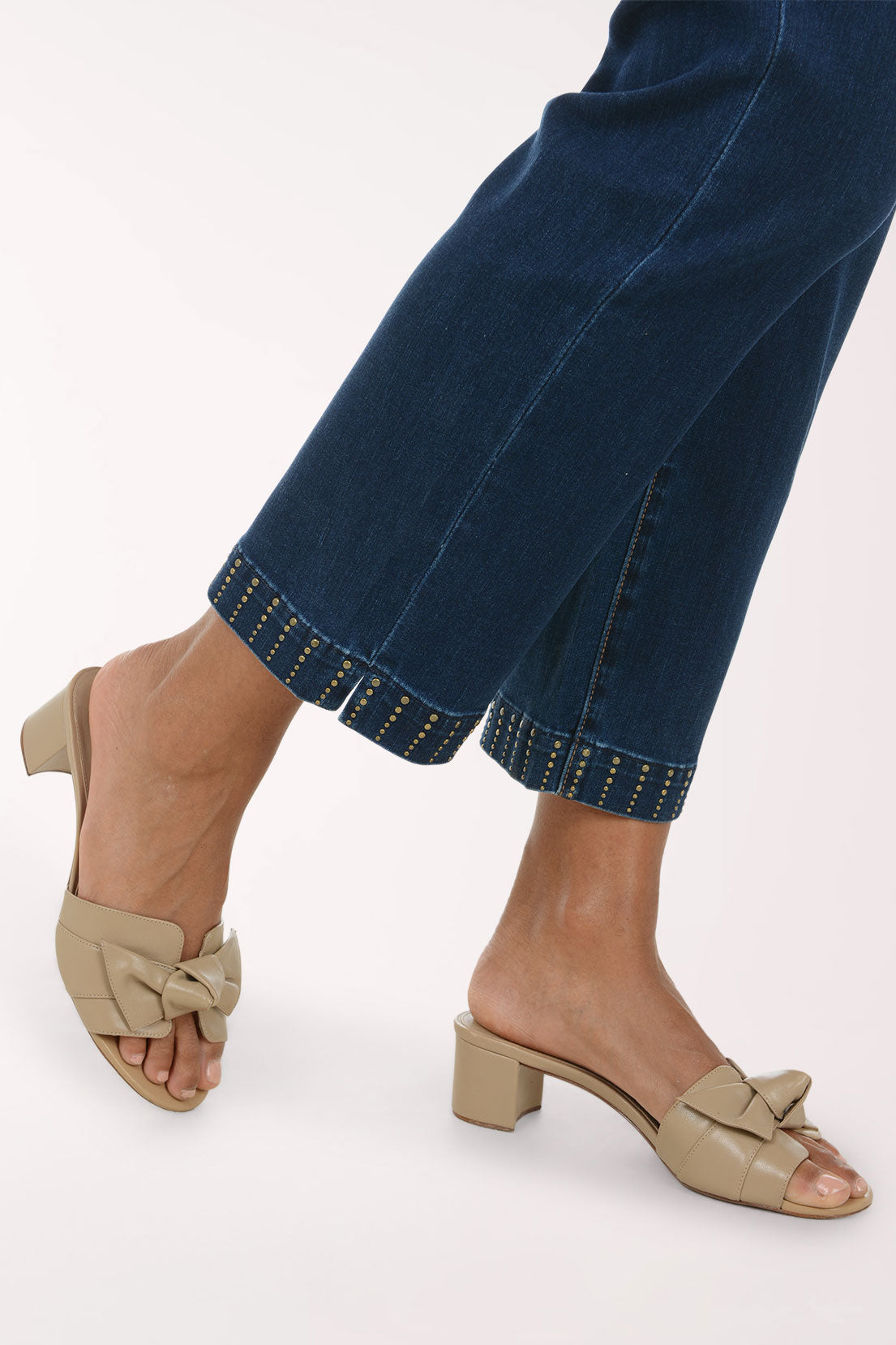Beige open-toe mule sandals with a knotted bow detail on the strap and a low block heel, shown on a model wearing dark blue jeans with gold stud trim at the hem.