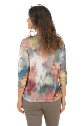 Woman wearing a scoop neck 3/4 sleeve colorful tie-dye shirt with arms crossed on a white background. Back shot.