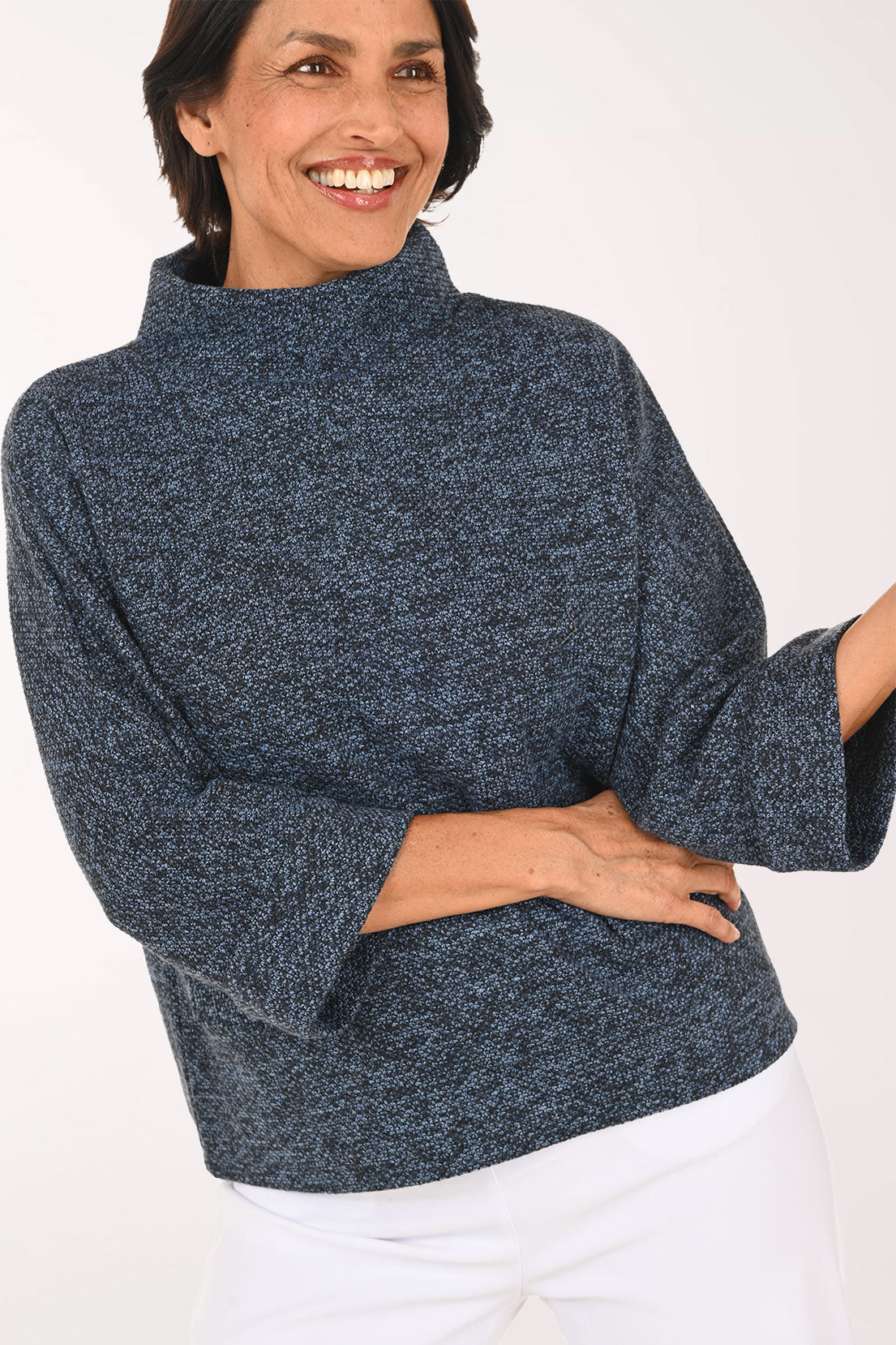 A smiling woman wearing a heathered navy blue mock-neck knit sweater with three-quarter bell sleeves, paired with white pants, against a plain white background.