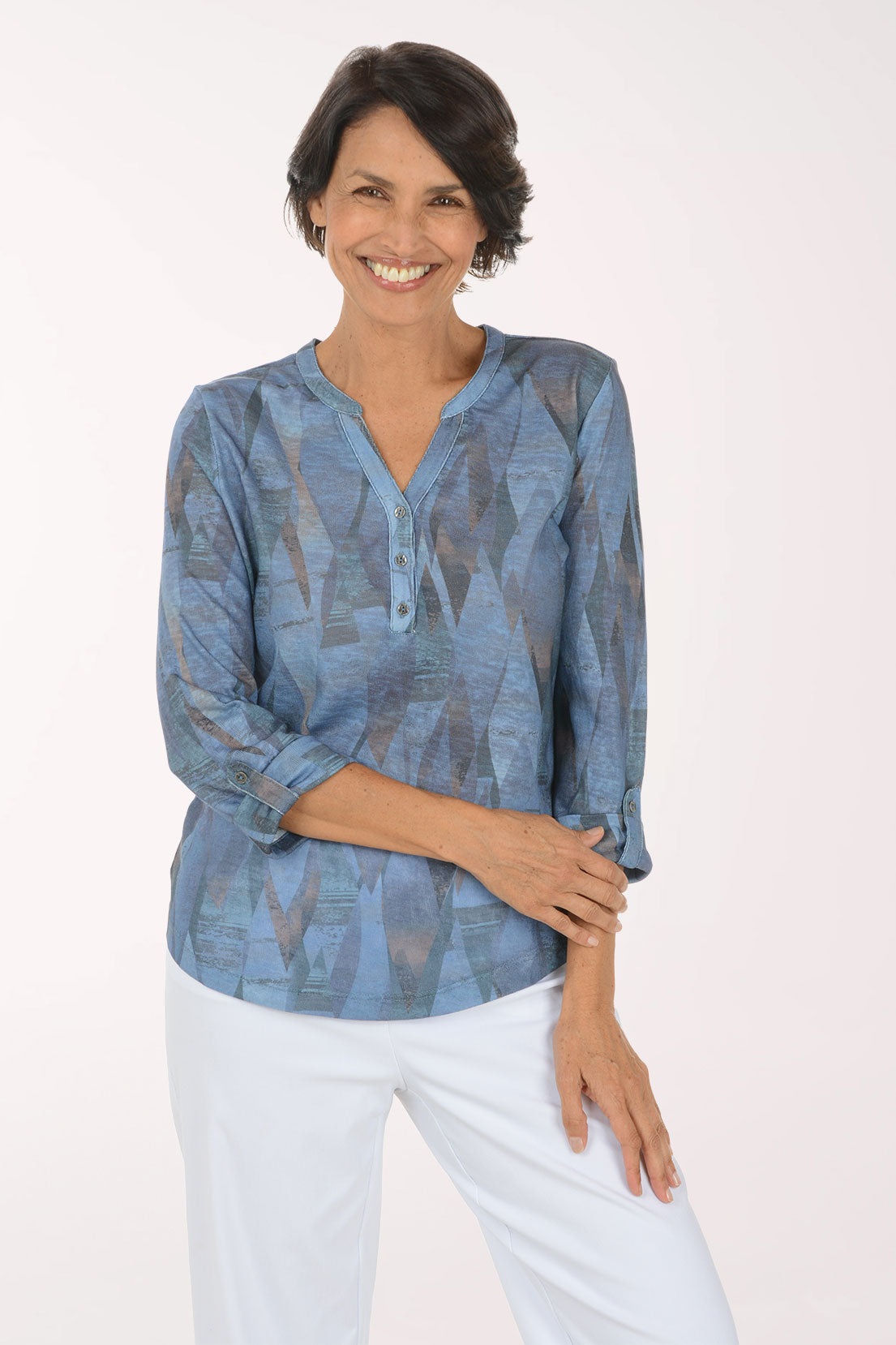 A smiling woman with short dark hair models a blue patterned long-sleeve blouse with white pants against a plain light background.