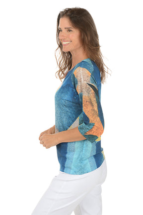 Woman wearing a colorful patterned top with white pants on a white background. Side shot.