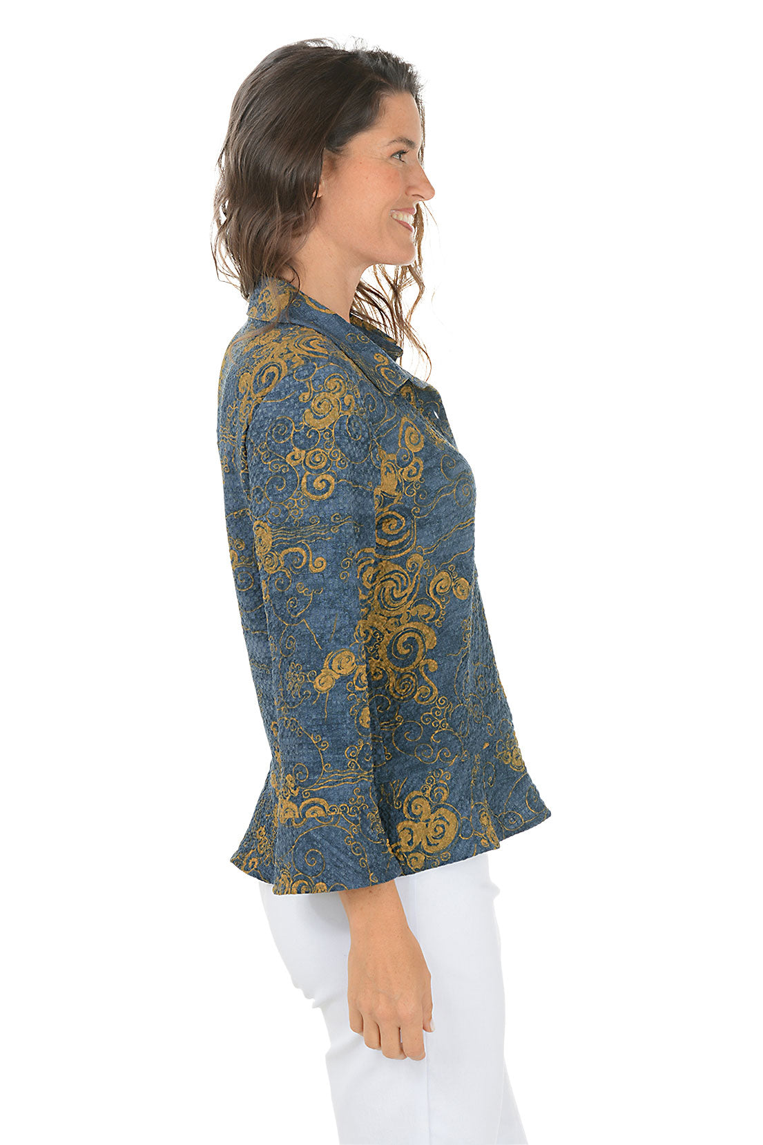 Woman wearing a blue and gold patterned blouse with white pants on a white background. Side shot.