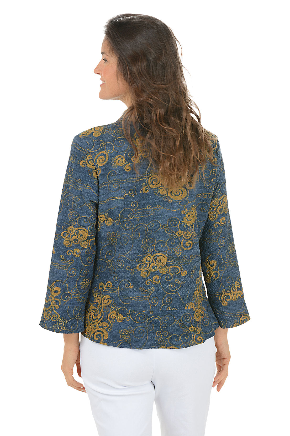 Woman wearing a blue and gold patterned blouse with white pants on a white background. Back shot.