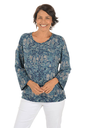 Woman wearing a blue paisley patterned top and white pants on a white background.