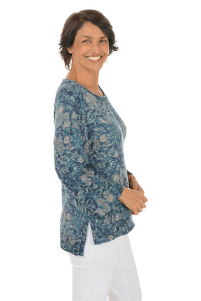 Woman wearing a blue paisley patterned top and white pants on a white background. Side shot.