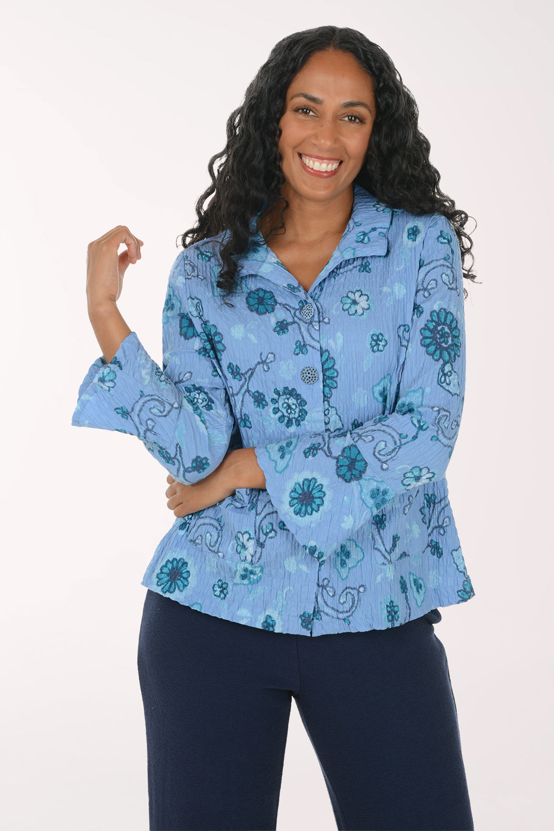 A smiling woman with long curly black hair models a light blue button-up blouse with a teal floral and paisley pattern, paired with dark navy pants, against a plain white studio background.