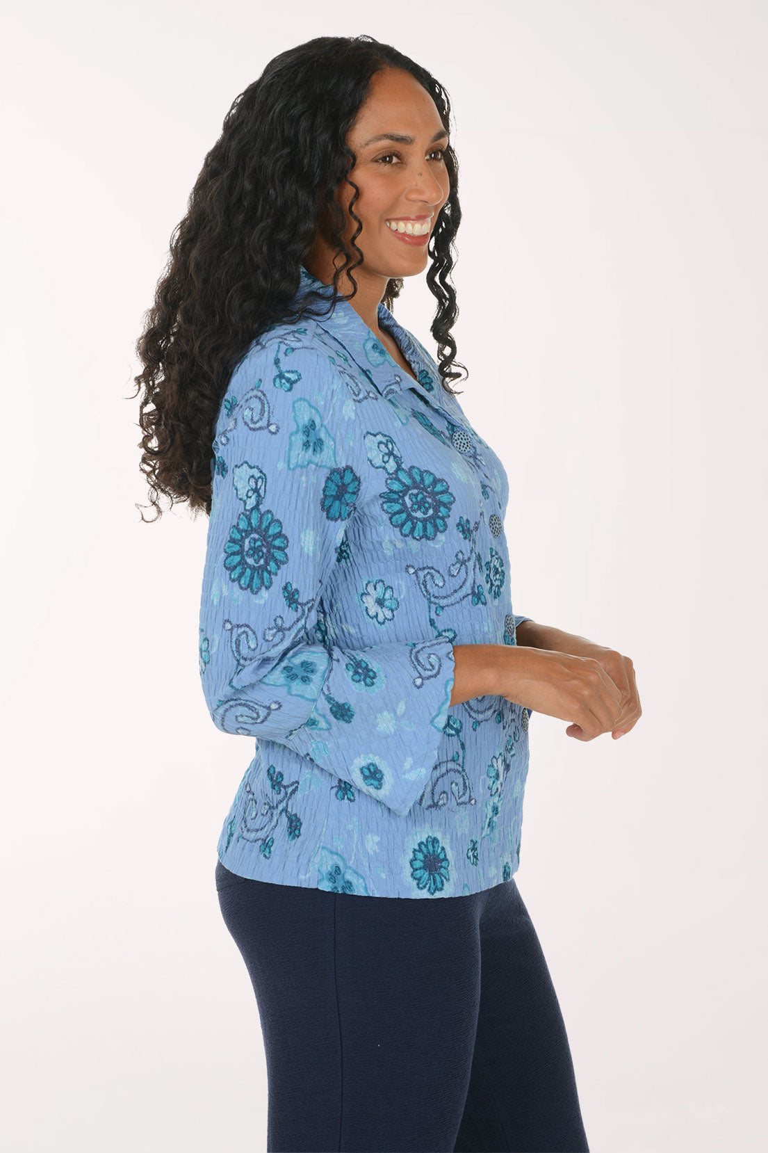 A smiling woman with long curly black hair models a light blue button-up blouse with a teal floral and paisley pattern, paired with dark navy pants, against a plain white studio background.