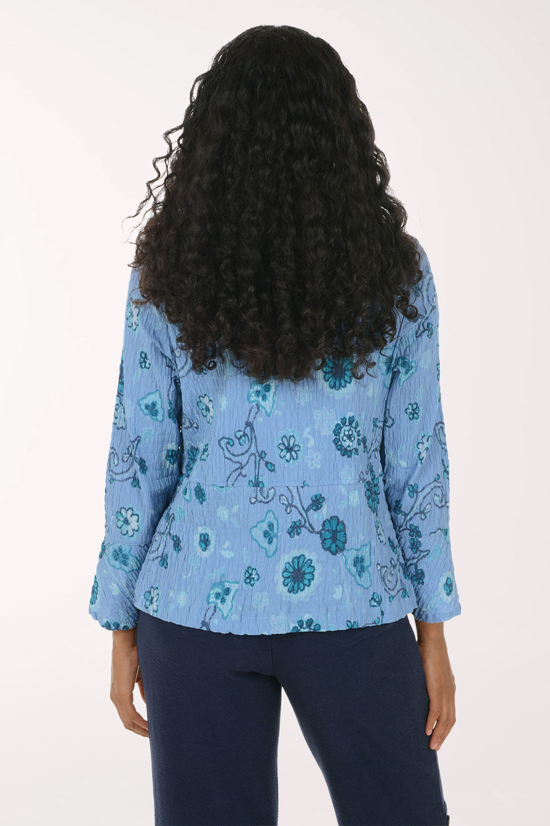A woman viewed from the back wearing a light blue long-sleeve blouse with a teal and dark blue floral pattern, paired with dark navy pants, against a plain white background.
