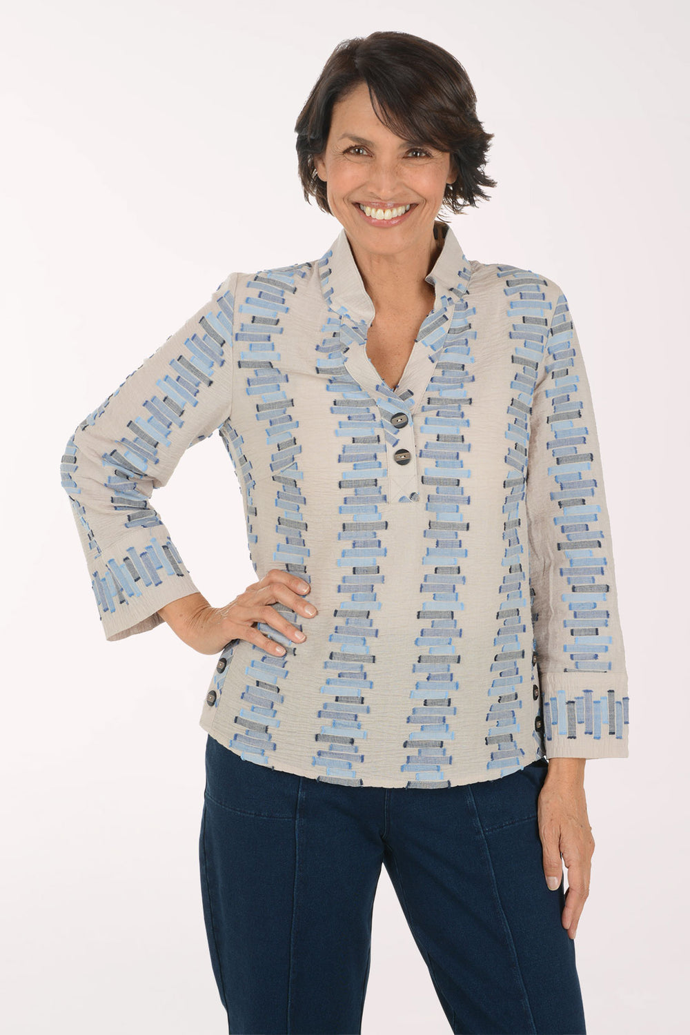A smiling woman wearing a light beige button-front blouse with a blue and gray geometric pattern, paired with dark blue pants, against a plain white studio background.