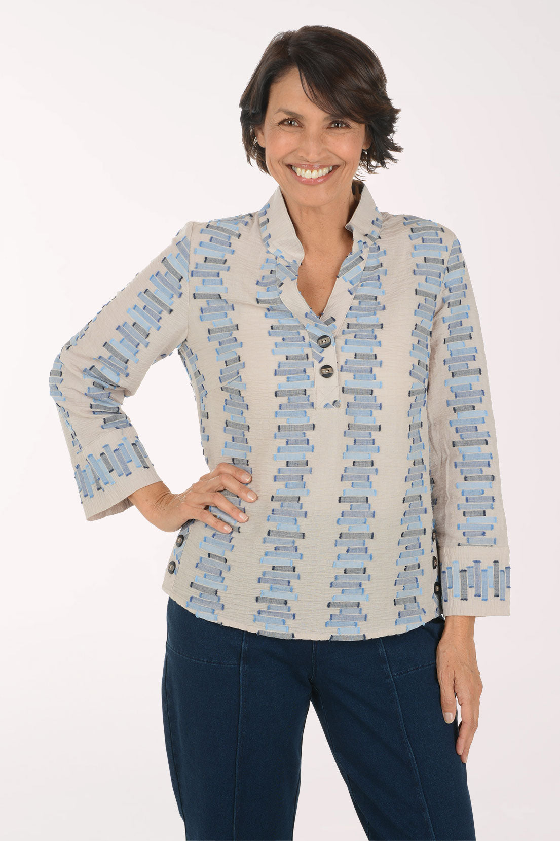 A smiling woman wearing a light beige button-front blouse with a blue and gray geometric pattern, paired with dark blue pants, against a plain white studio background.