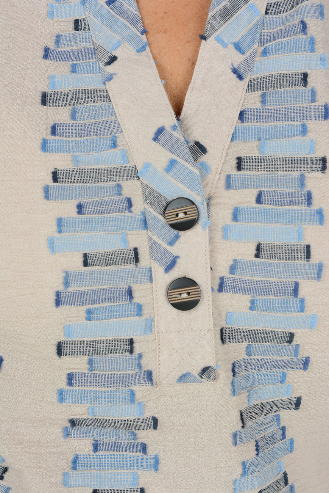 Close-up of a cream-colored textured top with a V-neckline and a decorative button placket. The fabric features vertical rows of light blue, medium blue, and navy striped rectangular patterns.