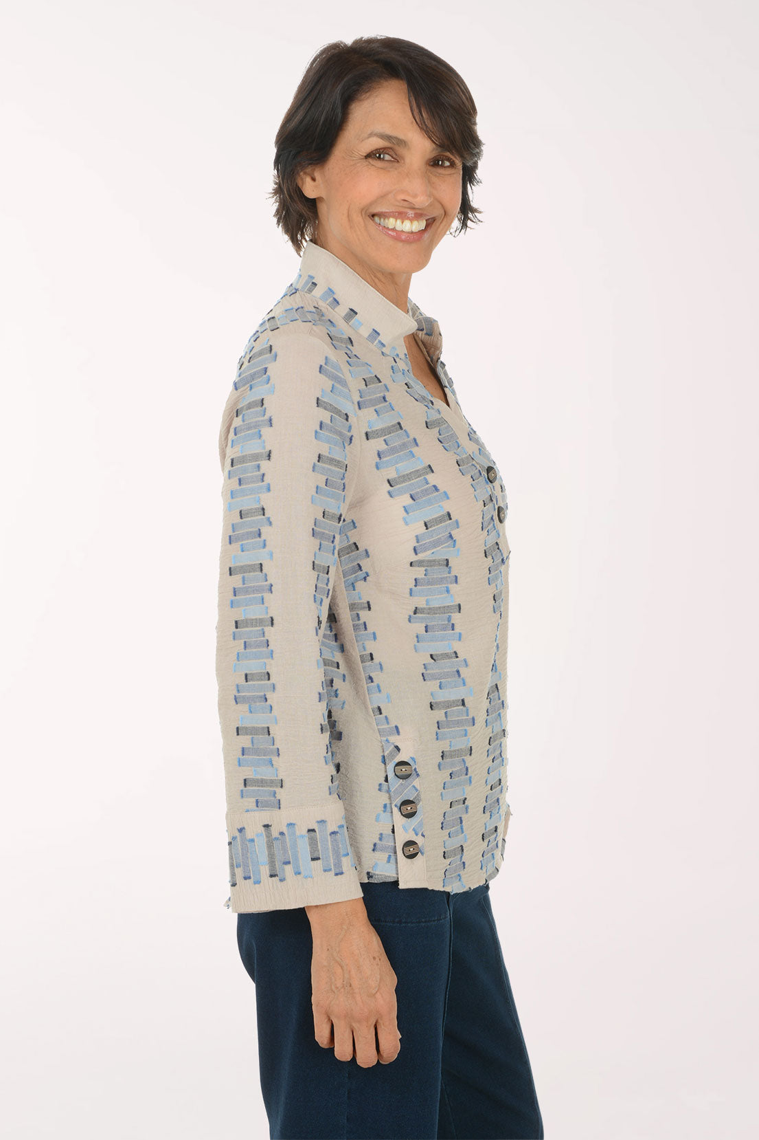 A smiling woman with short dark hair is shown in profile wearing a light beige blazer with a blue and gray geometric pattern, paired with dark blue pants, against a plain white studio background.