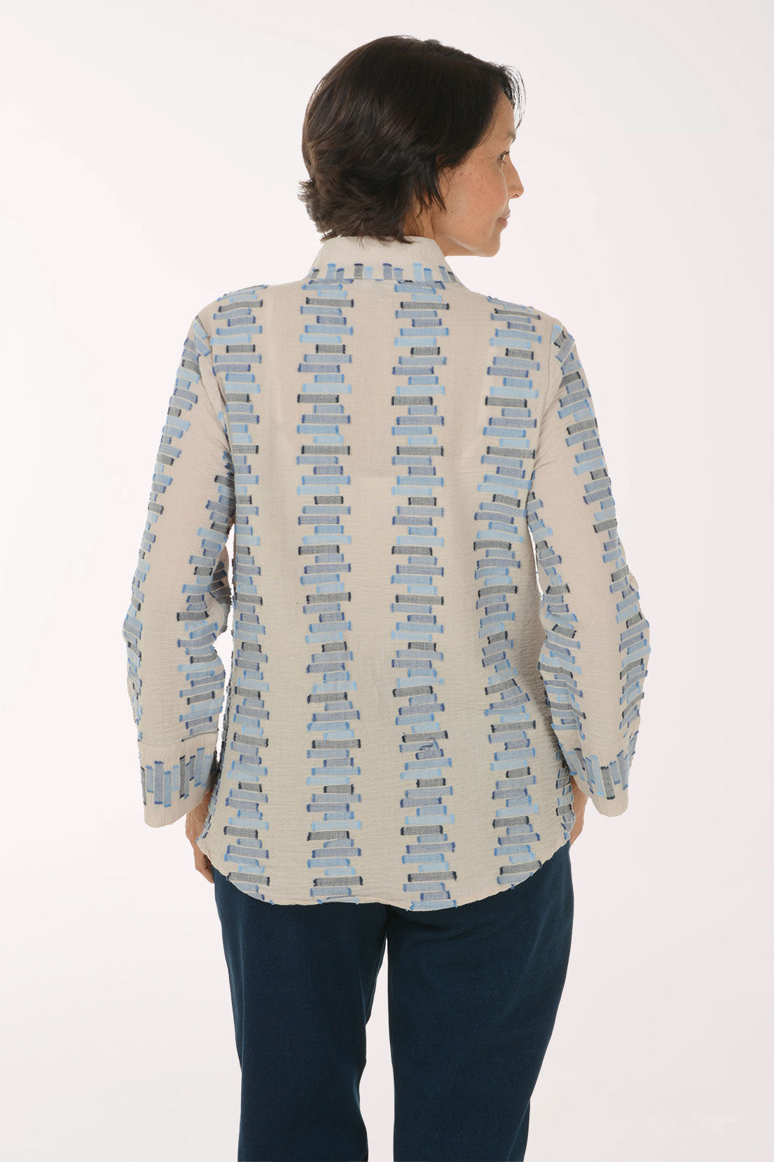 A woman shown from the back wearing a cream-colored long-sleeve button-up shirt with a repeating vertical stack-of-books pattern in light blue and gray, paired with dark blue pants, against a plain white studio background.