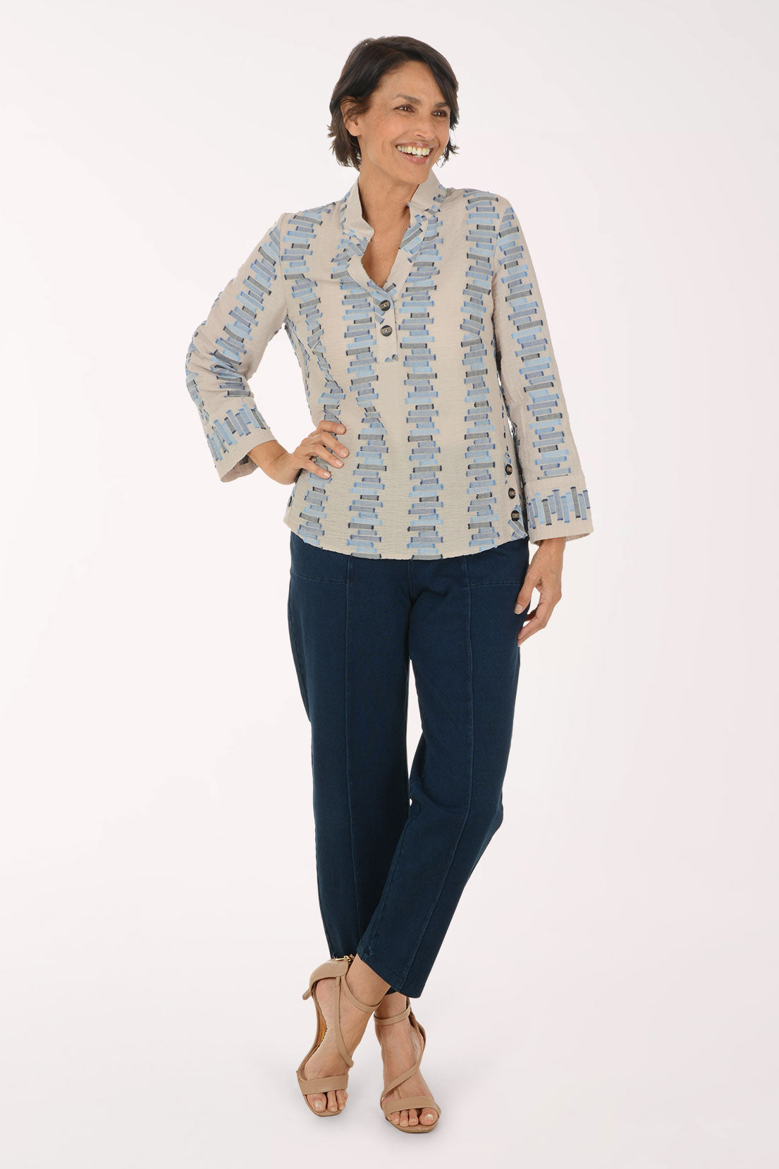 A woman modeling a light beige long-sleeve button-up shirt with a blue and gray geometric pattern, paired with navy blue pants and beige high-heeled sandals, against a plain white studio background.