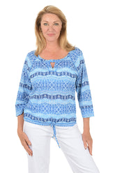 Woman wearing a blue patterned top and white pants on a white background.
