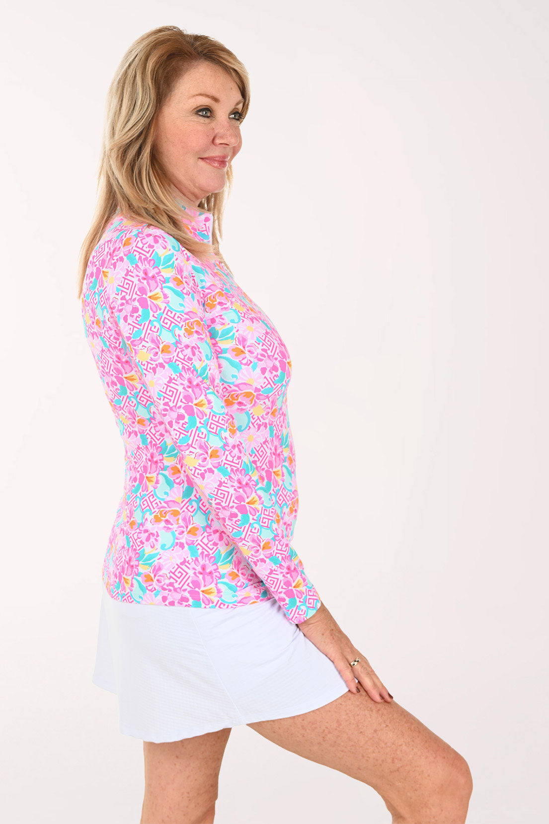 A woman modeling a long-sleeve collared top with a vibrant pink, blue, and orange floral pattern, paired with a white skirt, against a plain white studio background.