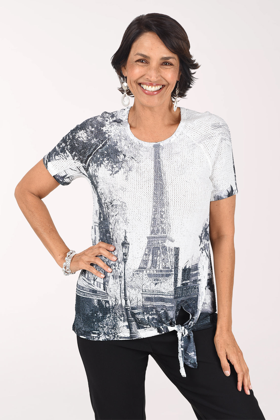 Front view of a woman in a white eyelet short raglan sleeve top printed with a black and grey Paris street scene and the Eiffel Tower, tied at the side of the hem, worn with black pants.