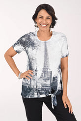 Front view of a woman in a white eyelet short raglan sleeve top printed with a black and grey Paris street scene and the Eiffel Tower, tied at the side of the hem, worn with black pants.