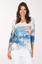 Smiling woman in a scoop neck 3/4 sleeve top printed with an ocean sunset scene of blue waves, a golden sky, white palm fronds and seashells, with a high-low hem, worn with white pants