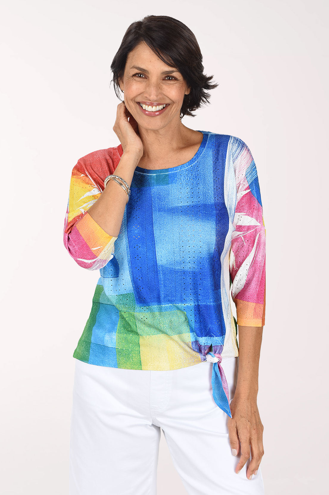 Smiling woman with a hand at her neck in a scoop neck eyelet top color-blocked in cobalt, red, sunflower yellow, green and pink, worn with white pants
