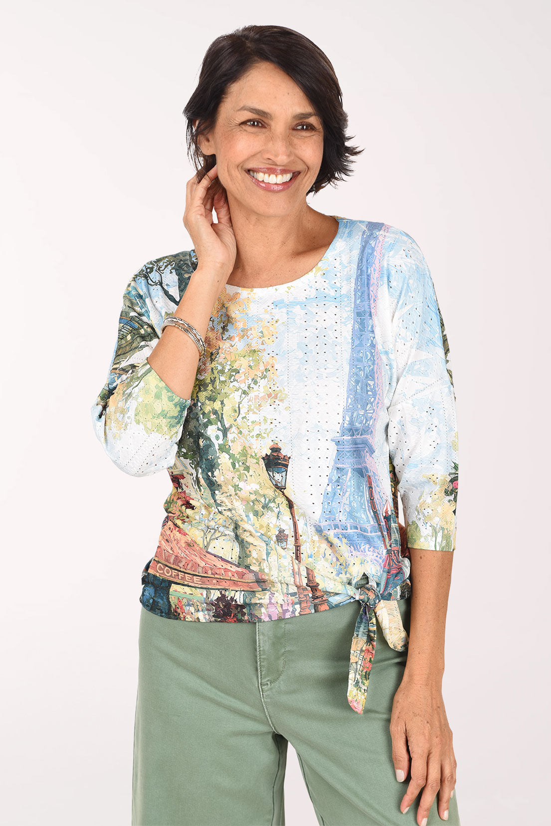 Smiling woman in a 3/4 dolman sleeve eyelet top printed with a painted Paris scene of the Eiffel Tower, flowering trees and a lamppost, tied at the front hem and worn with sage green pants