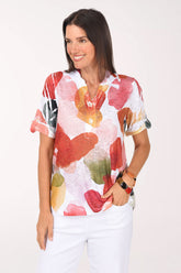 Model wearing the Impulse Drifting Petals Sequin Accented Polo Top, white with oversized red, coral and golden yellow petals, over white pants, front view