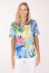 Model wearing the Impulse Colorful Abstract Sequin Accented Polo Top, a bright watercolor print in cobalt, yellow, coral and green, over white pants, front view