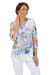 Woman wearing a colorful venice patterned 3/4 sleeve top with white pants on a white background.