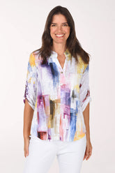 Smiling woman in a white roll sleeve top with a watercolor brushstroke print in violet, pink, saffron and sky blue, a sequined mandarin collar and split V-neck placket, worn with white pants