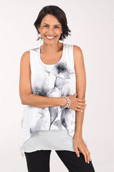 Front view of a woman in a white sleeveless scoop neck blouse with large soft grey watercolor flowers, a pointed shark bite hem layered over a white and grey inner tank, worn with black pants.